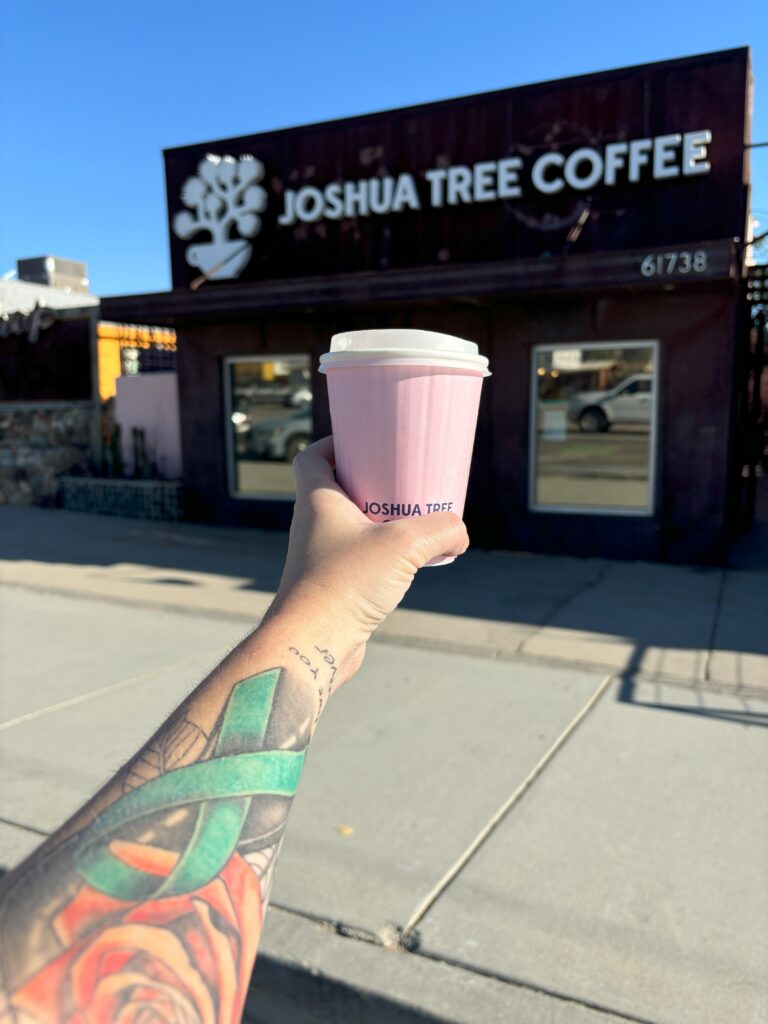 A hand holding a pink to go coffee cup in front of Joshua Tree Coffee